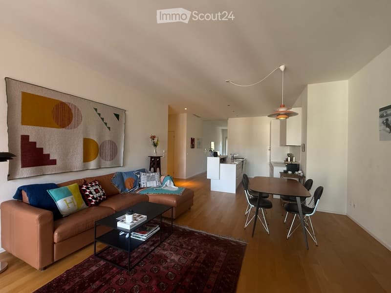 Modern living room with a brown leather sofa, colorful pillows, a coffee table with books and a cup, a dining table with chairs, a kitchen area with a white island, and a large abstract tapestry on the wall.