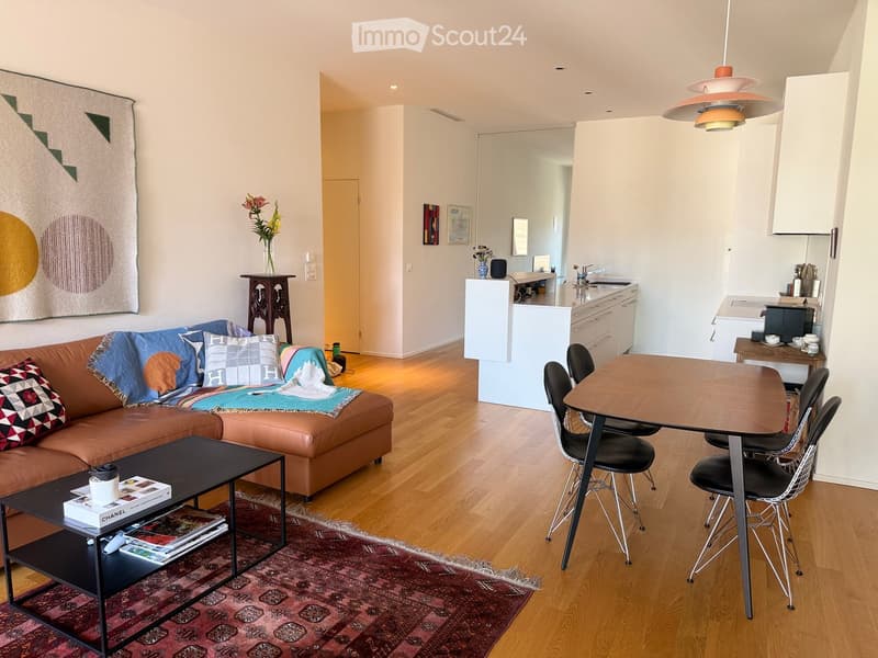 A modern, open-concept living and dining area with a leather sofa, coffee table, dining table with chairs, and a small kitchen area. The space features wooden flooring, a colorful rug, and decorative items including a vase with flowers and framed artwork.