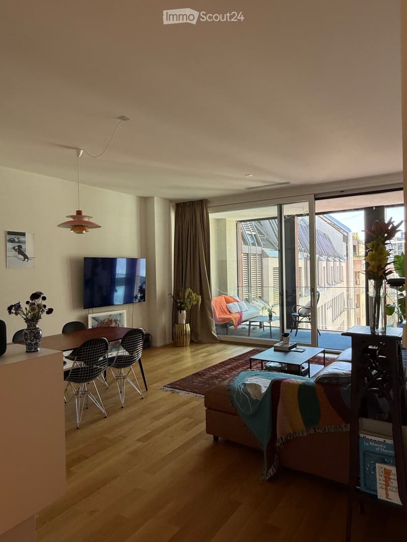 Modern living room with wooden flooring, large sliding glass doors leading to a balcony, sofa, coffee table, dining table with chairs, and a television mounted on the wall.