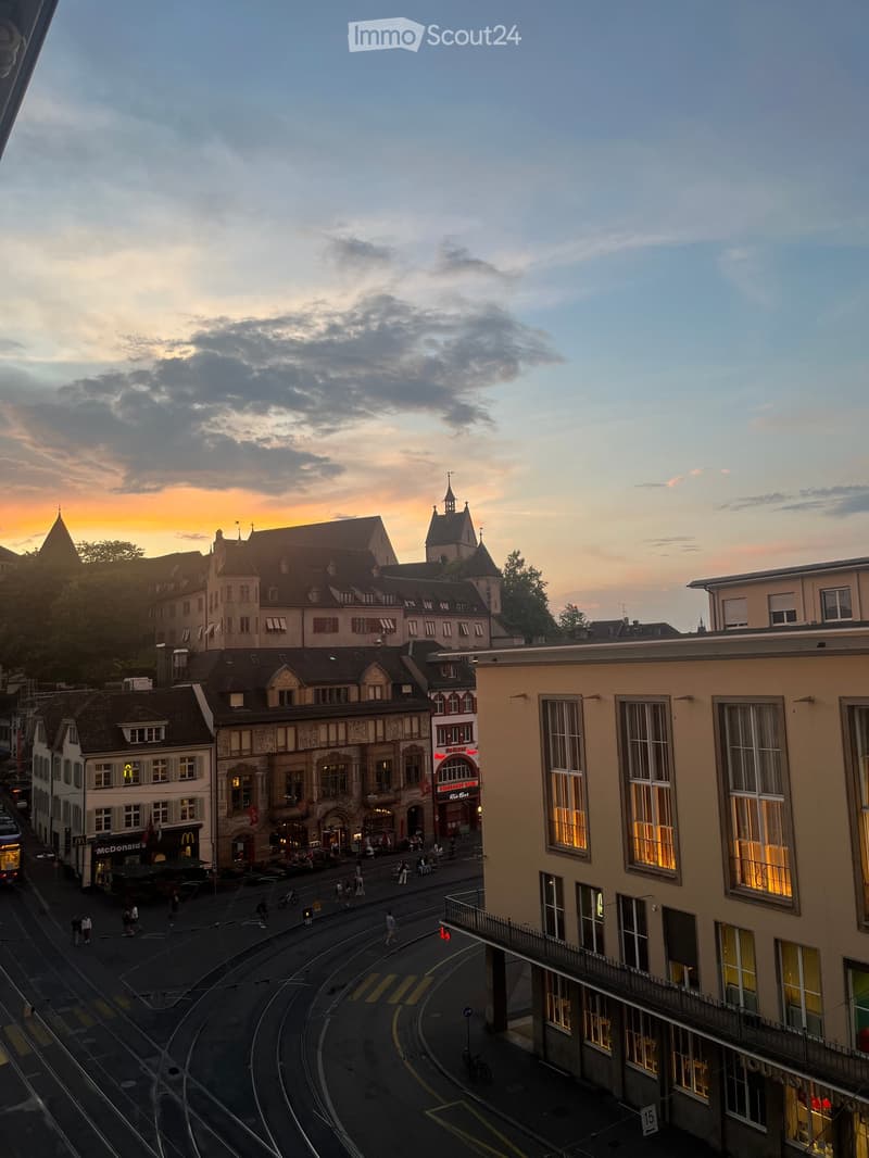 A cityscape view at sunset showing a mix of historic and modern buildings, with a tram line running through the street and people walking along the sidewalk.