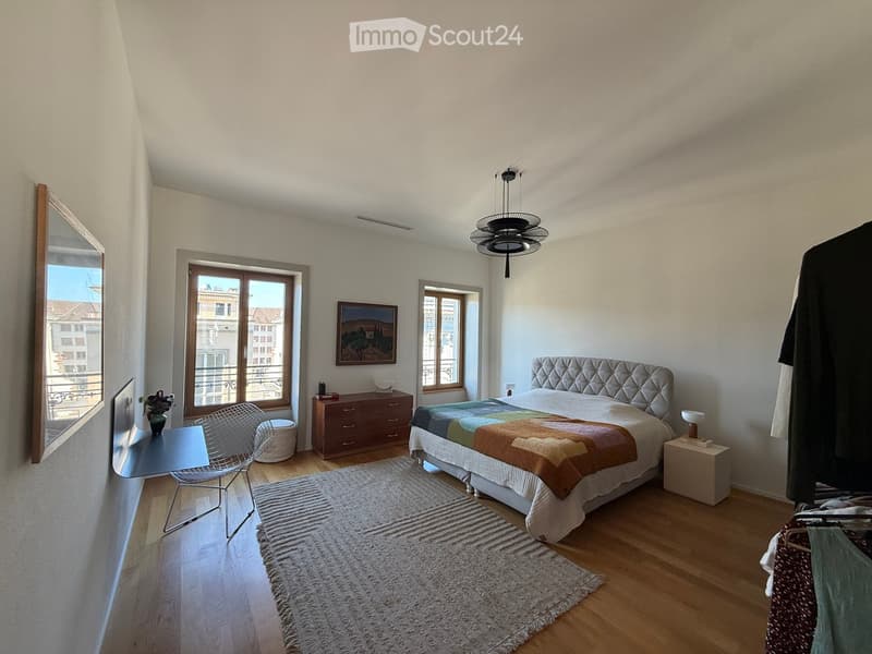 Modern bedroom with a large bed, nightstands, a chair, a small table, a mirror, and a picture frame on the wall. The room features wooden flooring and a large window with a view of the city.