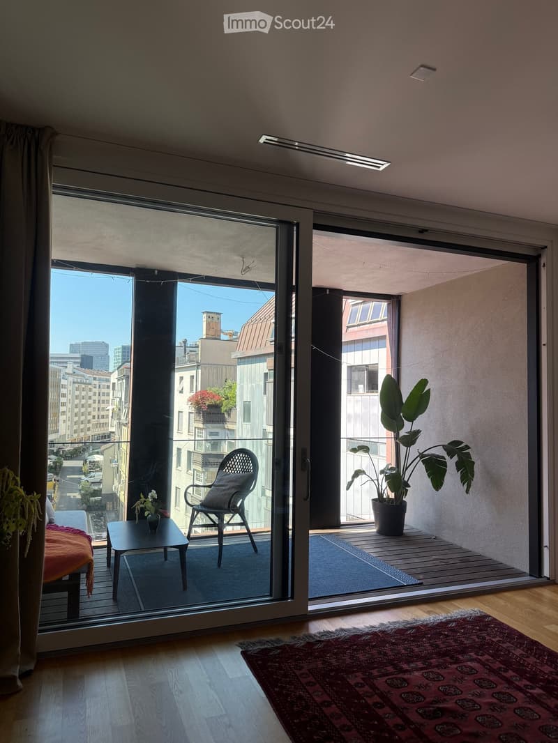 Modern living room with large sliding glass doors opening to a balcony, featuring a sofa, coffee table, chair, and potted plants.