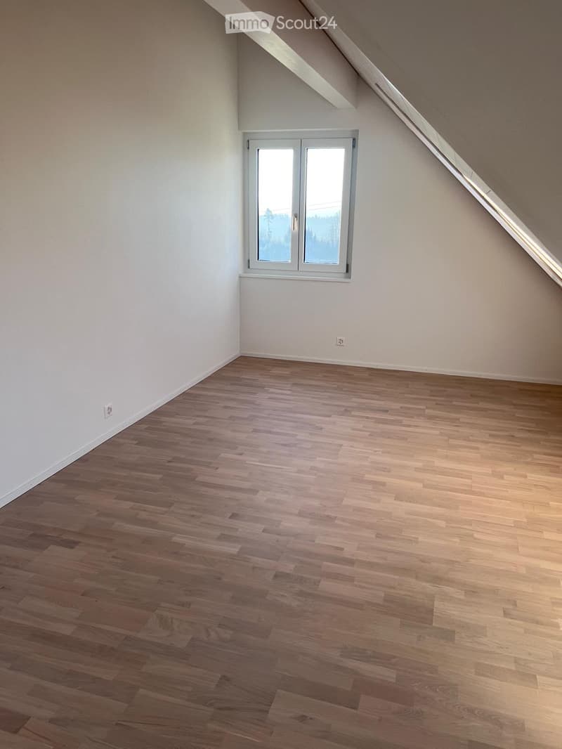Empty room with wooden flooring and white walls, featuring a large window with a view of trees.