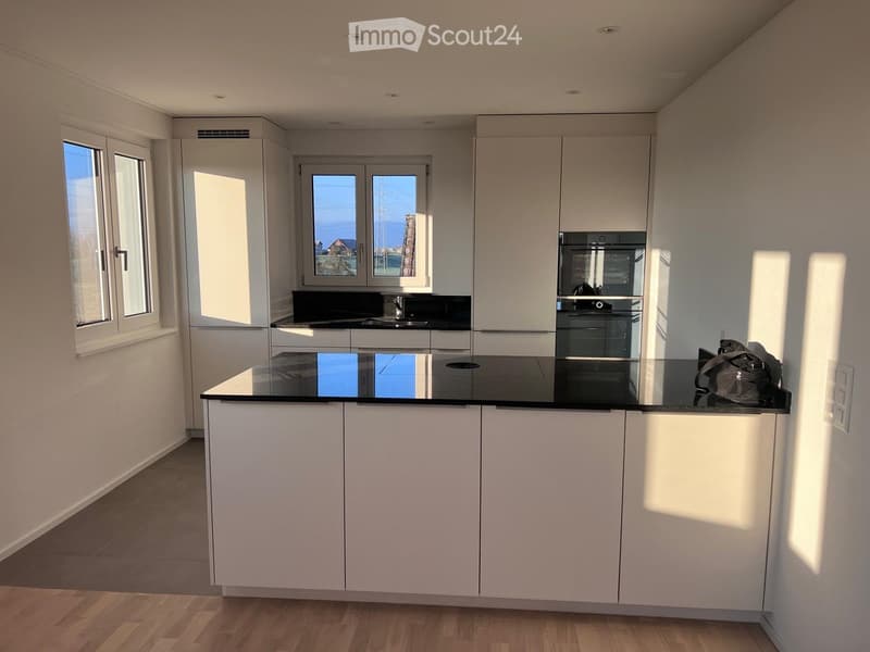 Modern kitchen with white cabinets, black countertops, and a central island. Features include a sink, an oven, and large windows letting in natural light.