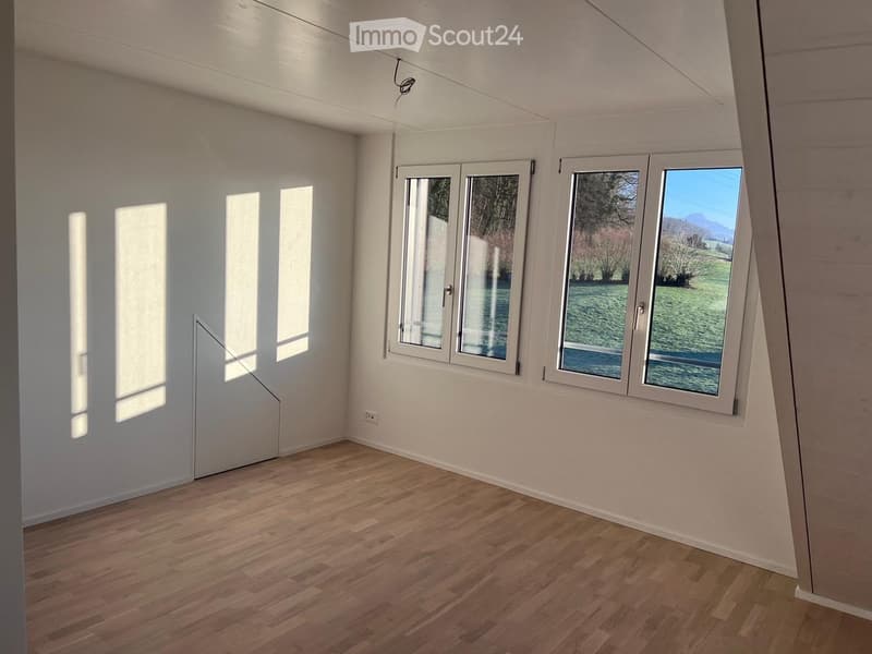 Empty room with wooden flooring, white walls, and large windows offering a view of a green landscape.