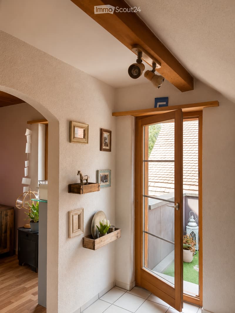A narrow hallway with wooden beams on the ceiling, white walls, and a wooden door leading to an outdoor area. Shelves with decorative items and framed pictures are mounted on the wall. The floor is a combination of wooden planks and white tiles.