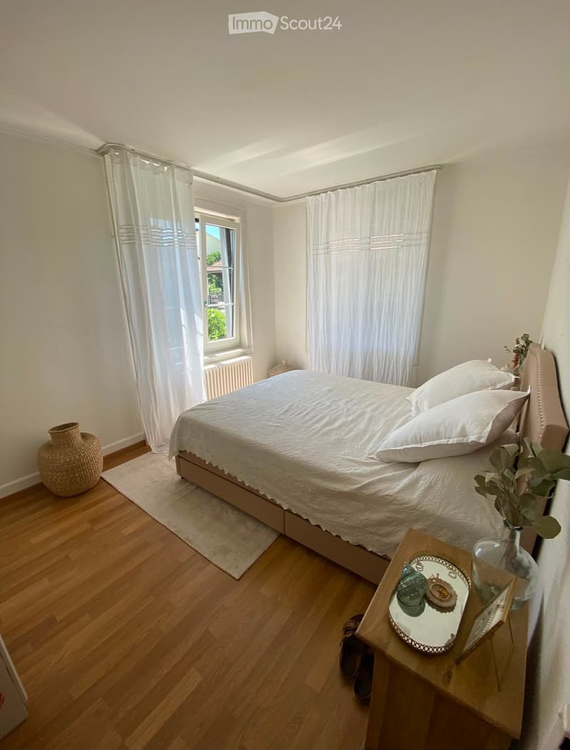 Cozy bedroom with wooden flooring, a neatly made bed with white bedding, a nightstand with a mirror, decorative items, and a wicker basket near the window.