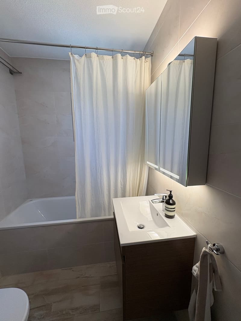Modern bathroom with a white sink, dark wooden cabinet, bathtub, mirror, and a shower curtain. A towel hangs on a rack, and a bottle of soap is placed on the sink.