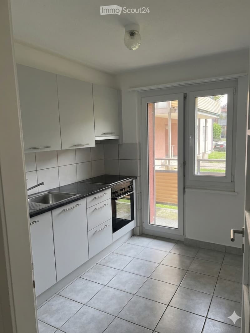 Modern kitchen with white cabinets, stainless steel sink, oven, and tiled floor. Features a large window and a sliding glass door leading to an outdoor area.