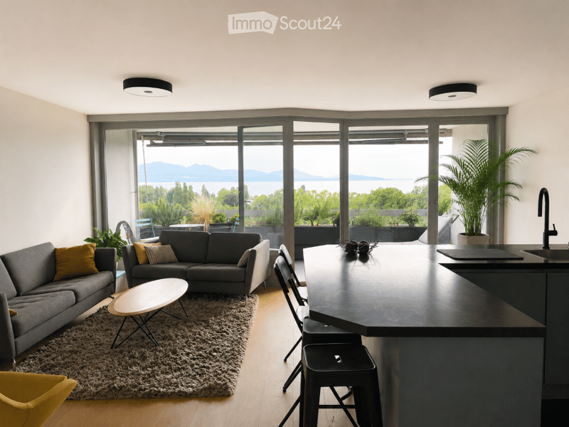 Modern open-plan living and dining area with large windows offering a scenic view, featuring a grey sectional sofa, coffee table, dining chairs, kitchen island, and plants.