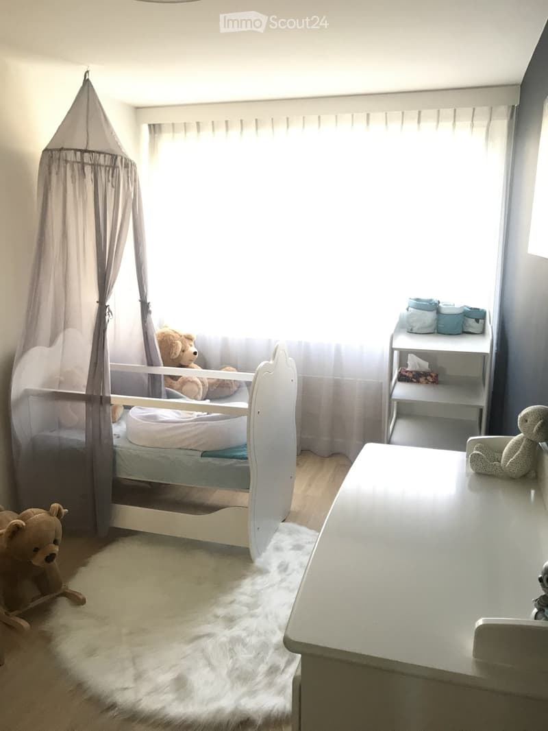 A well-lit children's bedroom with a white baby bed, a small table, a shelf with baby items, and several stuffed toys.