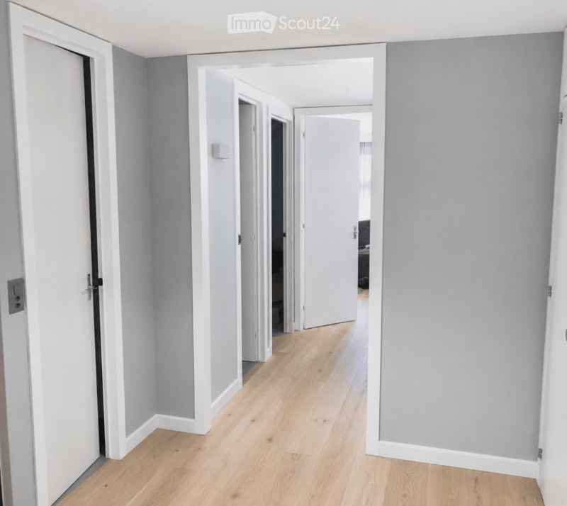 Empty hallway with light wooden flooring, grey walls, and white door frames. No furniture or equipment visible.