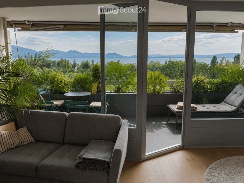 Living room with a grey sofa, wooden floor, large windows opening to a balcony with outdoor furniture and scenic mountain view.