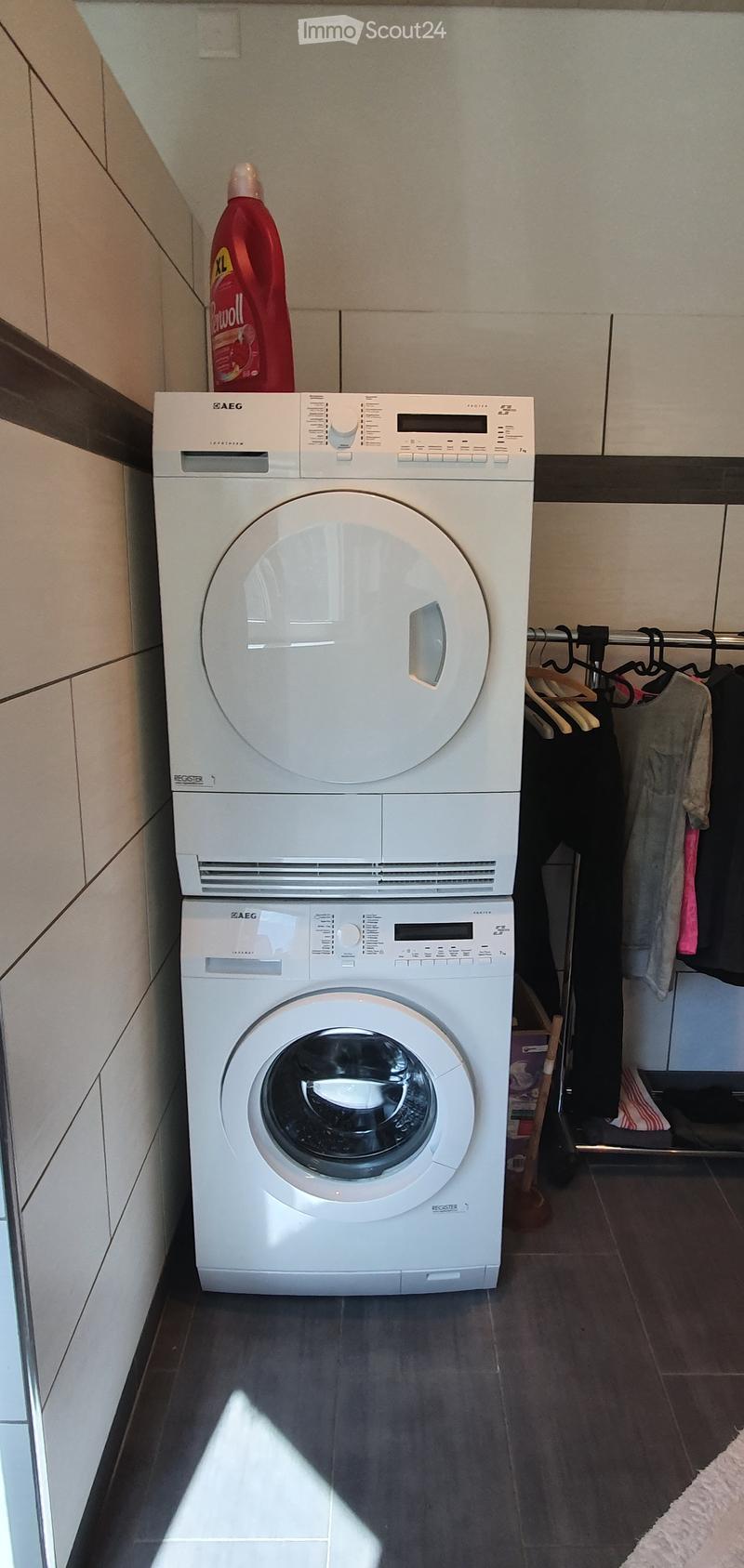 A compact bathroom with a stacked washing machine and dryer unit, a bottle of cleaning solution on top, and a clothes rack with hanging garments.