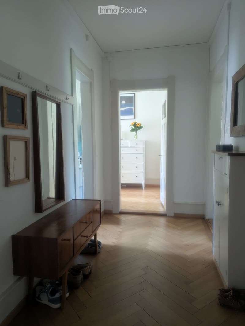 A narrow hallway with wooden flooring, white walls, and a wooden console table with a mirror. Shoes are placed near the console table, and an open doorway leads to another room with a dresser and a vase of flowers.
