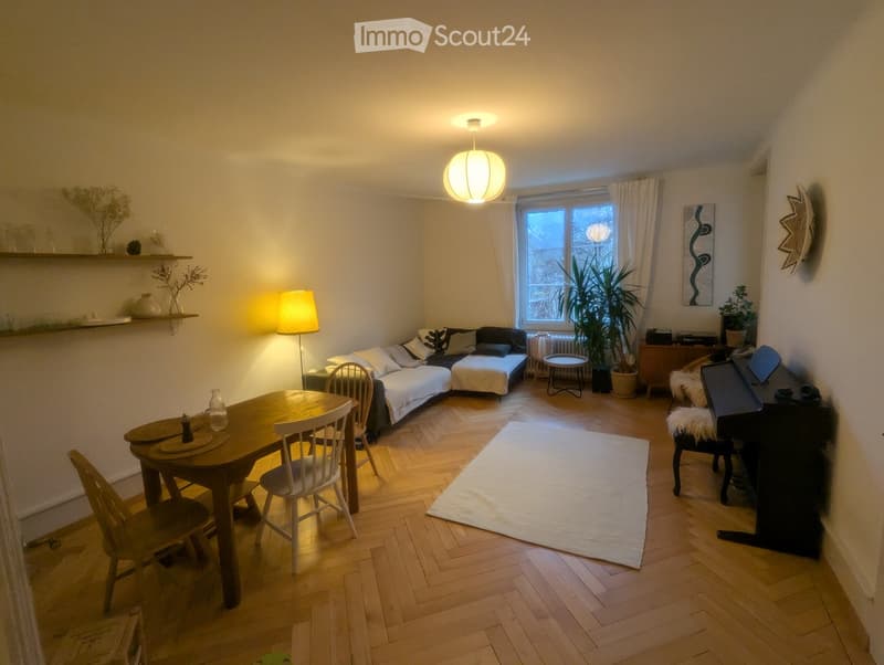 Cozy living room with wooden flooring, a sofa with pillows, a small table, chairs, a piano, a shelf with decorative items, and a large window with curtains.