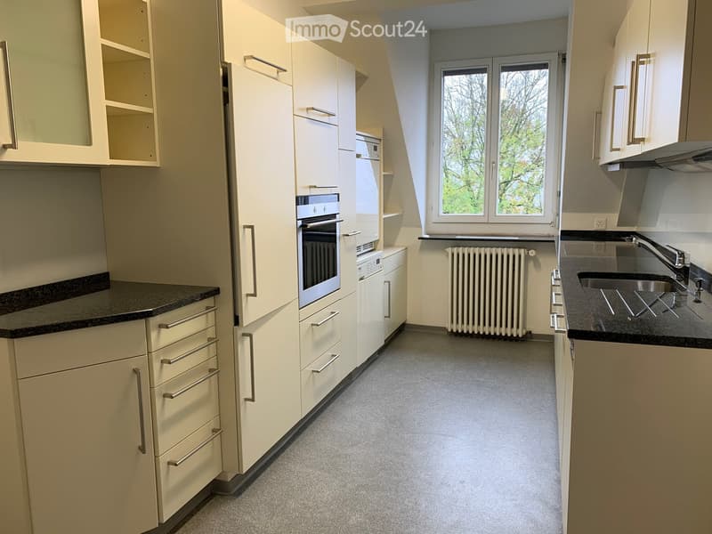 Modern kitchen with light-colored cabinets, stainless steel oven, sink, and radiator. Features a window with a view of trees.