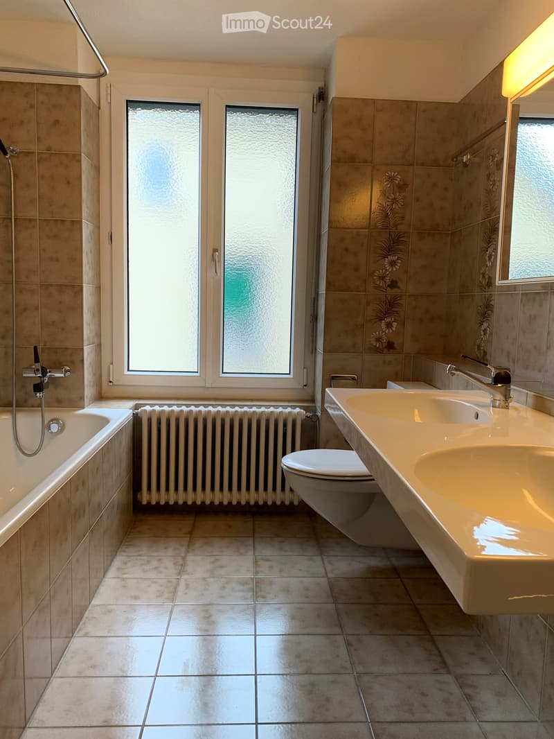 Modern bathroom with beige tiles, a bathtub, a toilet, a double sink, and a radiator. The room features large windows with frosted glass and a shower area with a glass door.