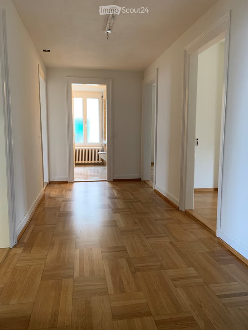 Empty hallway with wooden flooring, white walls, and recessed lighting. Doors lead to adjacent rooms, one with visible windows and a radiator.