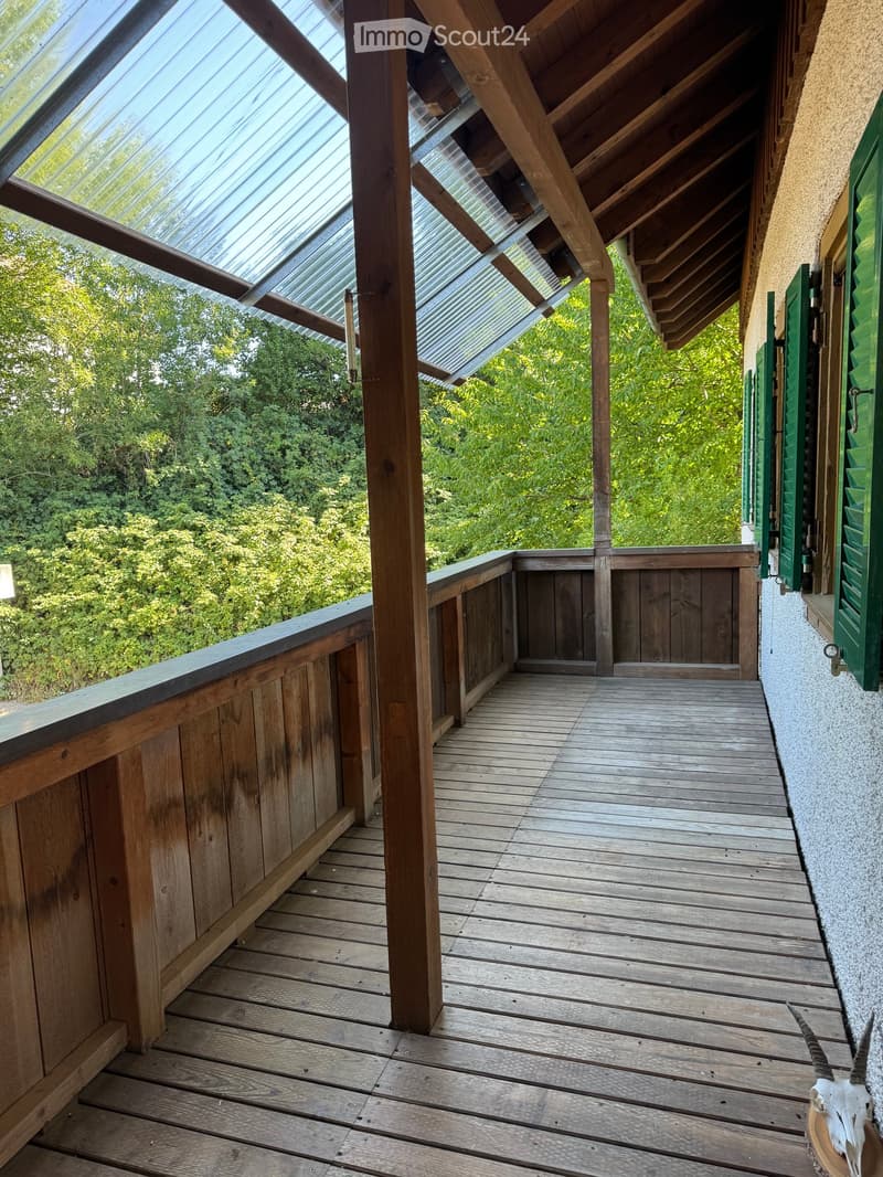 Wooden balcony with a slanted roof, wooden railing, and a view of greenery.