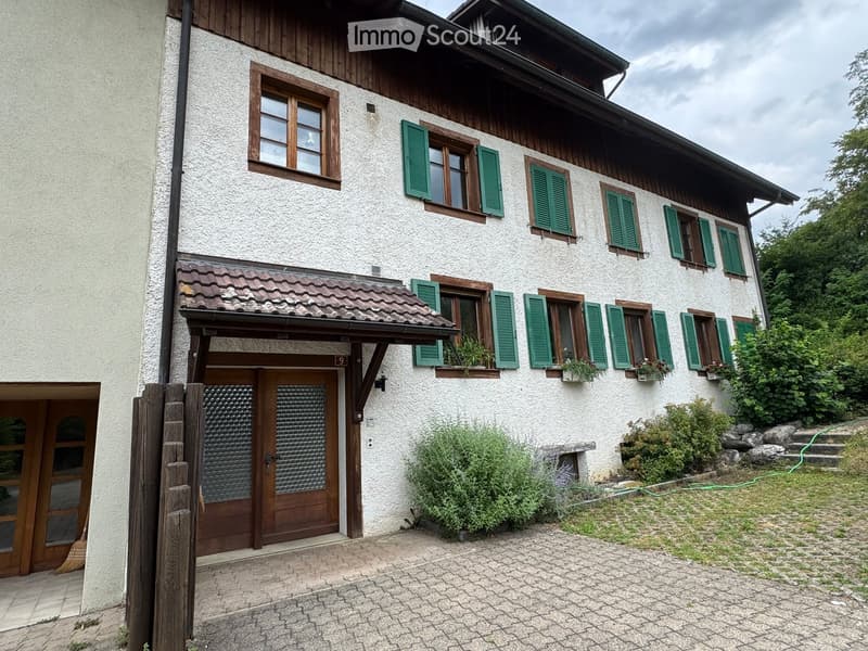 Two-story house with white textured walls, green shutters, wooden trim, and a tiled roof. Features a covered entrance with a wooden door and a small garden area with plants and a stone bench.