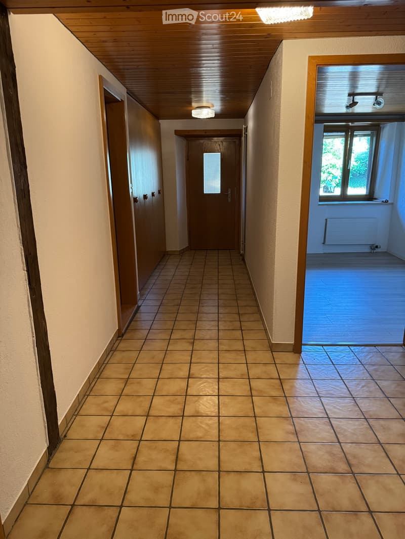 Empty hallway with tiled floor, wooden ceiling, and several doors leading to other rooms. Natural light from a window on the right side.