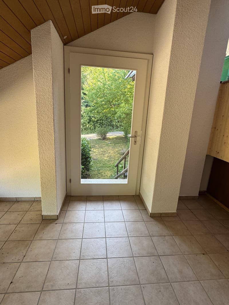 Empty hallway with tiled floor, beige walls, and a door leading to an outdoor area with greenery visible through the glass.