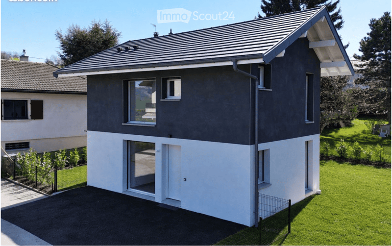 Modern two-story house with dark grey upper facade and white lower section, featuring large windows and a flat roof with a small dormer. The house is surrounded by a well-maintained lawn and small garden area.