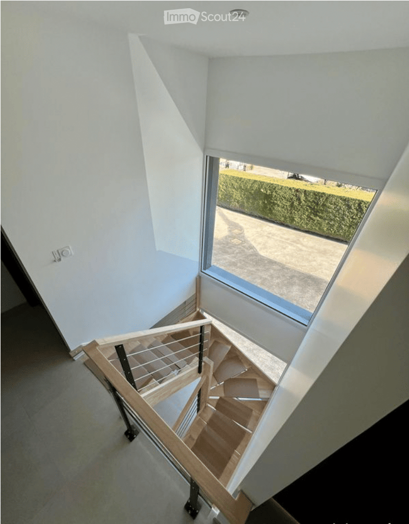 Modern staircase with wooden steps and metal railing, leading to an upper level. The walls are painted white, and there is a large window at the top providing natural light and a view of the garden outside.