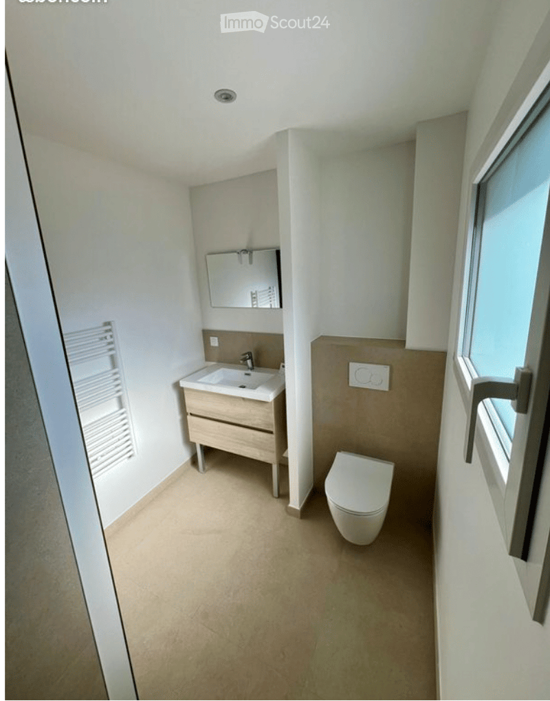 Modern bathroom with a white sink, wooden cabinet, mirror, and toilet. Light-colored walls and floor, with a window on the right side.
