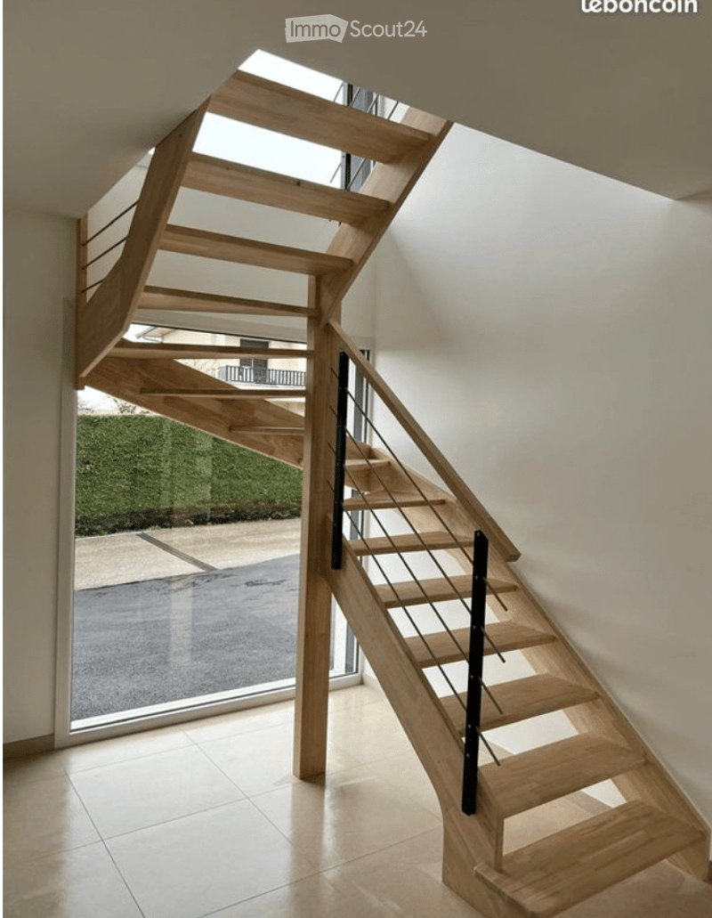 Spiral staircase with wooden steps and a modern metal railing, leading to an upper level. The staircase is illuminated by natural light from a skylight above.