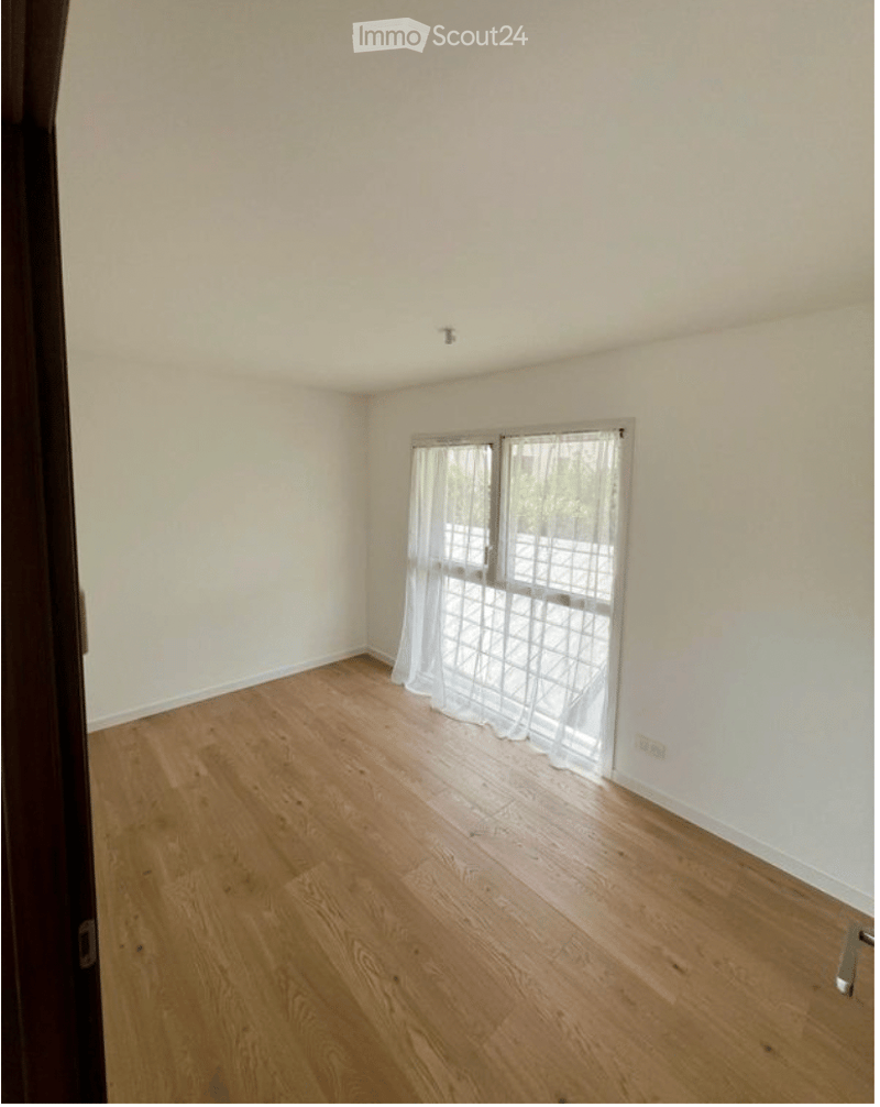 Empty room with wooden flooring, white walls, and large windows with sheer curtains.