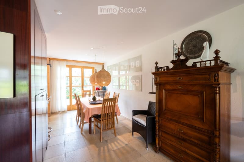 Dining area with a round wooden dining table, six chairs, a wicker armchair, a tall wooden cabinet, and decorative items on top. Natural light streams in from large windows with white curtains.