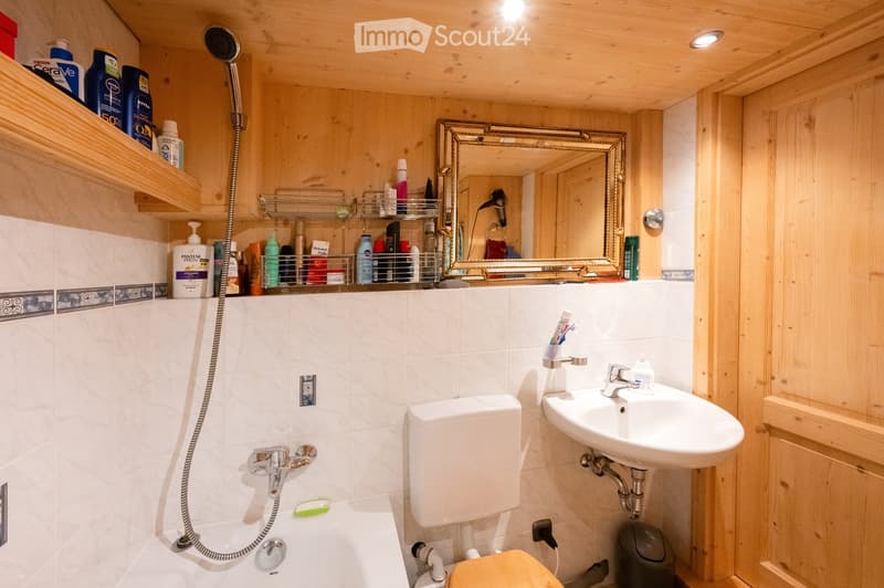 Cozy bathroom with wooden walls, white tiles, a bathtub, toilet, sink, mirror, and shelves holding toiletries.