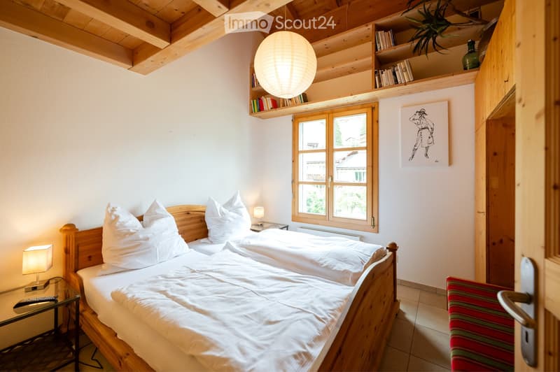 Cozy bedroom with two single beds, wooden headboards, nightstands with table lamps, a large window, built-in shelves with books and a decorative plant, and a colorful striped stool near the door.