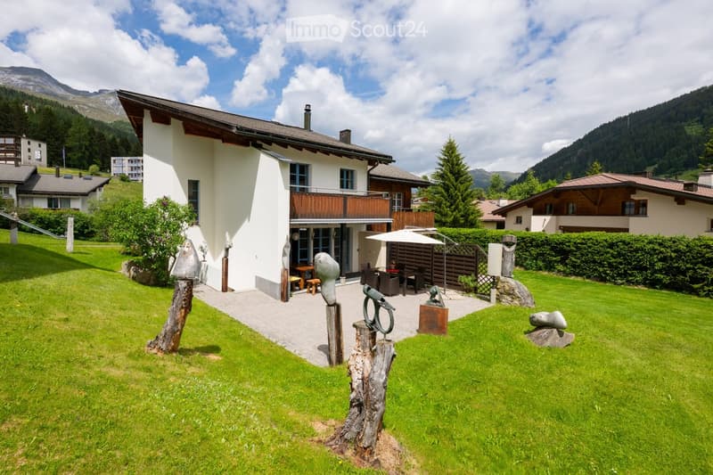 A two-story house with a balcony, surrounded by a well-maintained lawn and sculptures in the garden. The house features a modern design with large windows and a wooden balcony.