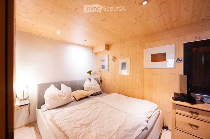 Cozy bedroom with wooden walls and ceiling, a double bed with white bedding and pillows, a nightstand with a lamp, a screen, and several picture frames on the wall.