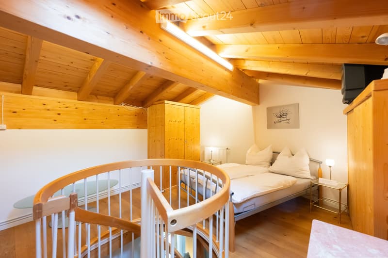 Cozy attic bedroom with wooden beams, slanted ceiling, and exposed wooden beams. Features a double bed with white bedding, a nightstand with a lamp, a wardrobe, and a small round table with a glass top.