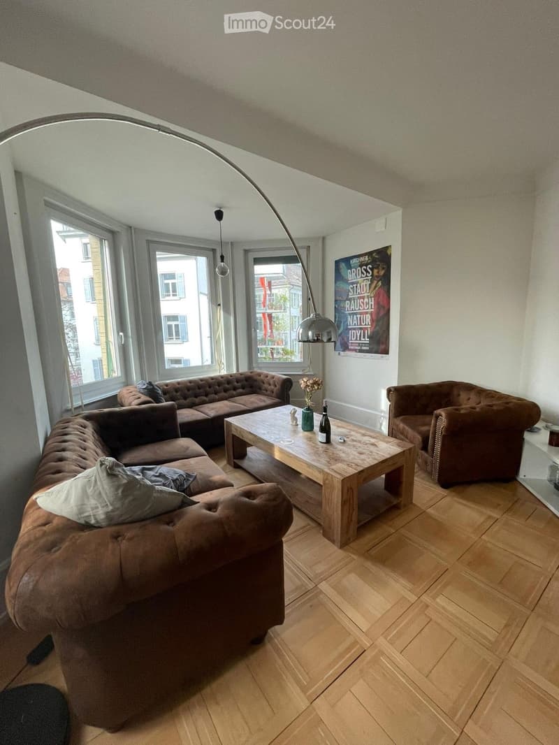 Modern living room with wooden flooring, large windows, a brown leather sofa, a wooden coffee table, and an armchair. A floor lamp and a vase with flowers are also present.