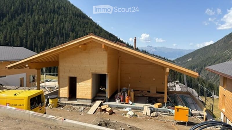 A newly constructed wooden house with a sloped roof and large windows, situated in a mountainous area with visible construction materials and tools around the base.