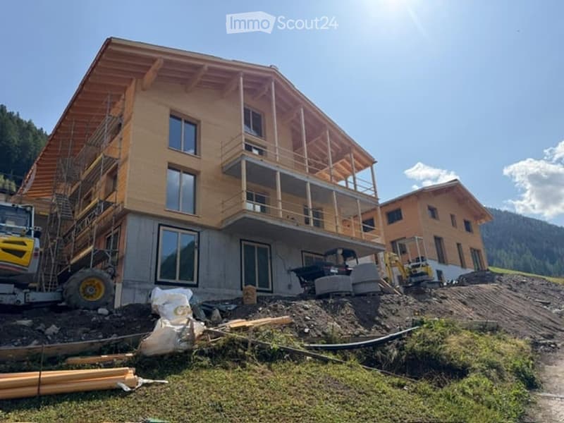 A two-story wooden house under construction with large windows and a raised ground floor, surrounded by construction materials and machinery.