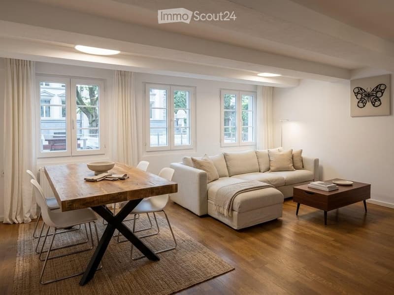 Modern living room with a light-colored sectional sofa, coffee table, dining table with chairs, and large windows with curtains. The room features a wooden floor and a neutral color palette.