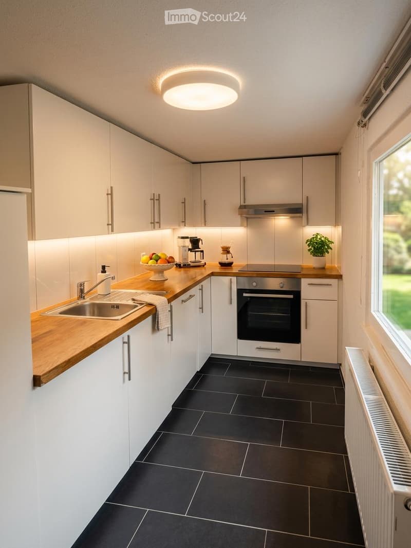 Modern kitchen with white cabinets, wooden countertops, black tiled floor, stainless steel sink, oven, coffee maker, fruit bowl, and small plant on the counter.