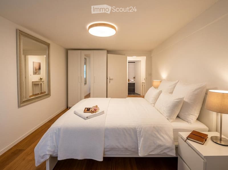 Modern, well-lit bedroom with a large bed, nightstand, lamp, wardrobe, and a mirror on the wall.