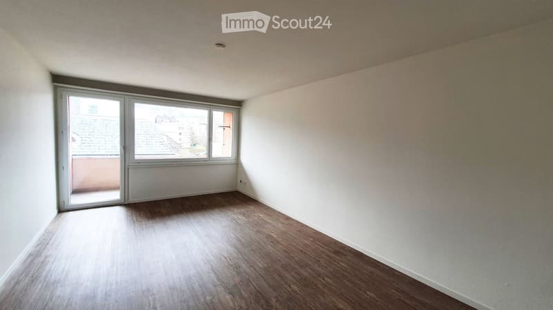 Empty room with wooden flooring, white walls, and large windows with a sliding door leading to a balcony.