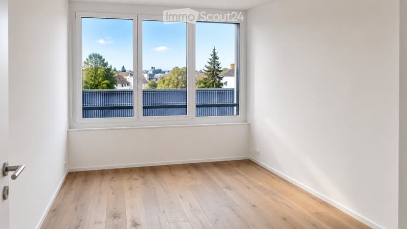 Empty room with light wooden flooring, white walls, and large windows offering a view of trees and buildings outside.