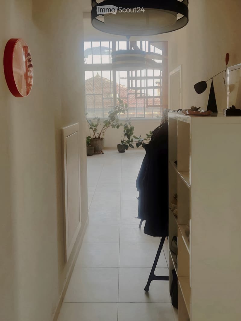A modern hallway with tiled flooring, light-colored walls, and a large window at the end. A coat is draped over a chair, and there are potted plants near the window. A decorative wall piece is visible on the left side.