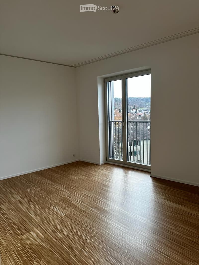 Empty room with wooden flooring, large sliding glass door leading to a balcony with a view of the city and surrounding landscape.