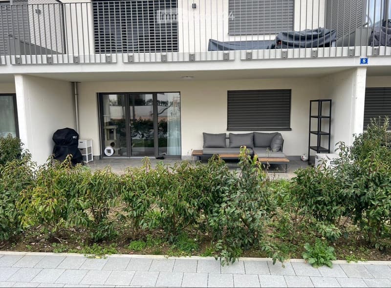 Outdoor patio area with a grey sofa, coffee table, shelving unit, and covered grill. The area is surrounded by greenery and has access to a balcony above.