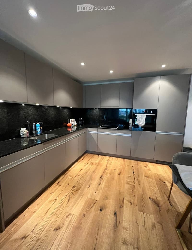 Modern kitchen with light grey cabinets, wooden flooring, and a black countertop. Features a sink, oven, and various small items on the counter.